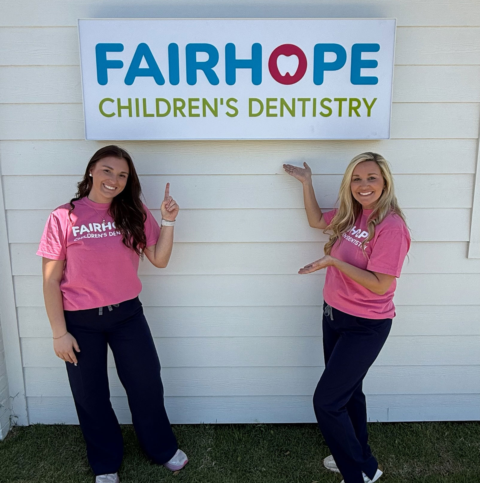 Two women in pink shirts pointing at the "Fairhope Children's Dentistry" sign on a building, promoting pediatric dental care services.