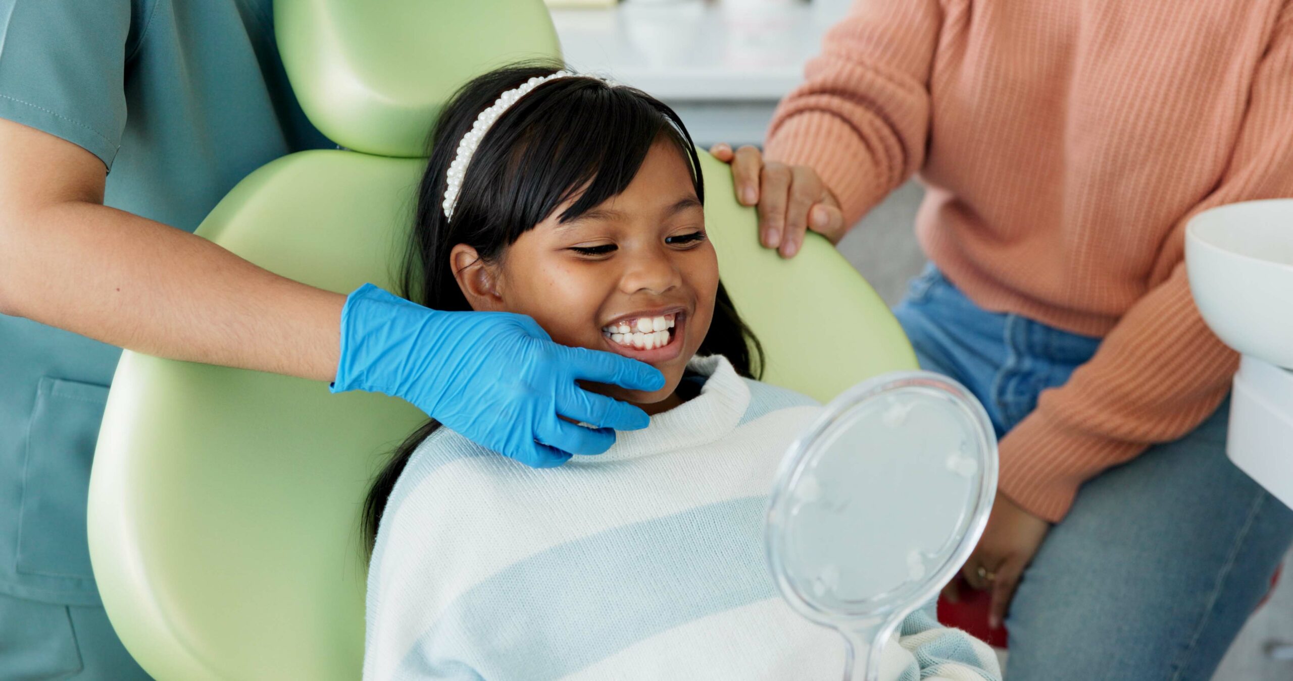 Child at pediatric dental appointment smiling while being examined by a dentist, with a parent supporting her, in a friendly dental office environment.
