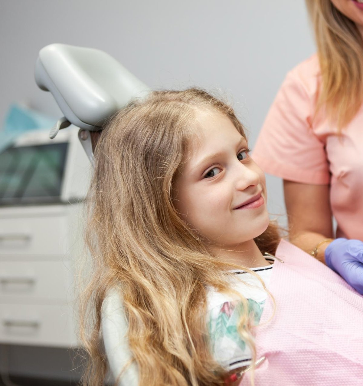 Child in dental chair smiling, wearing pink bib, with pediatric dentist in background, illustrating comfortable and friendly dental care environment at Fairhope Children's Dentistry.